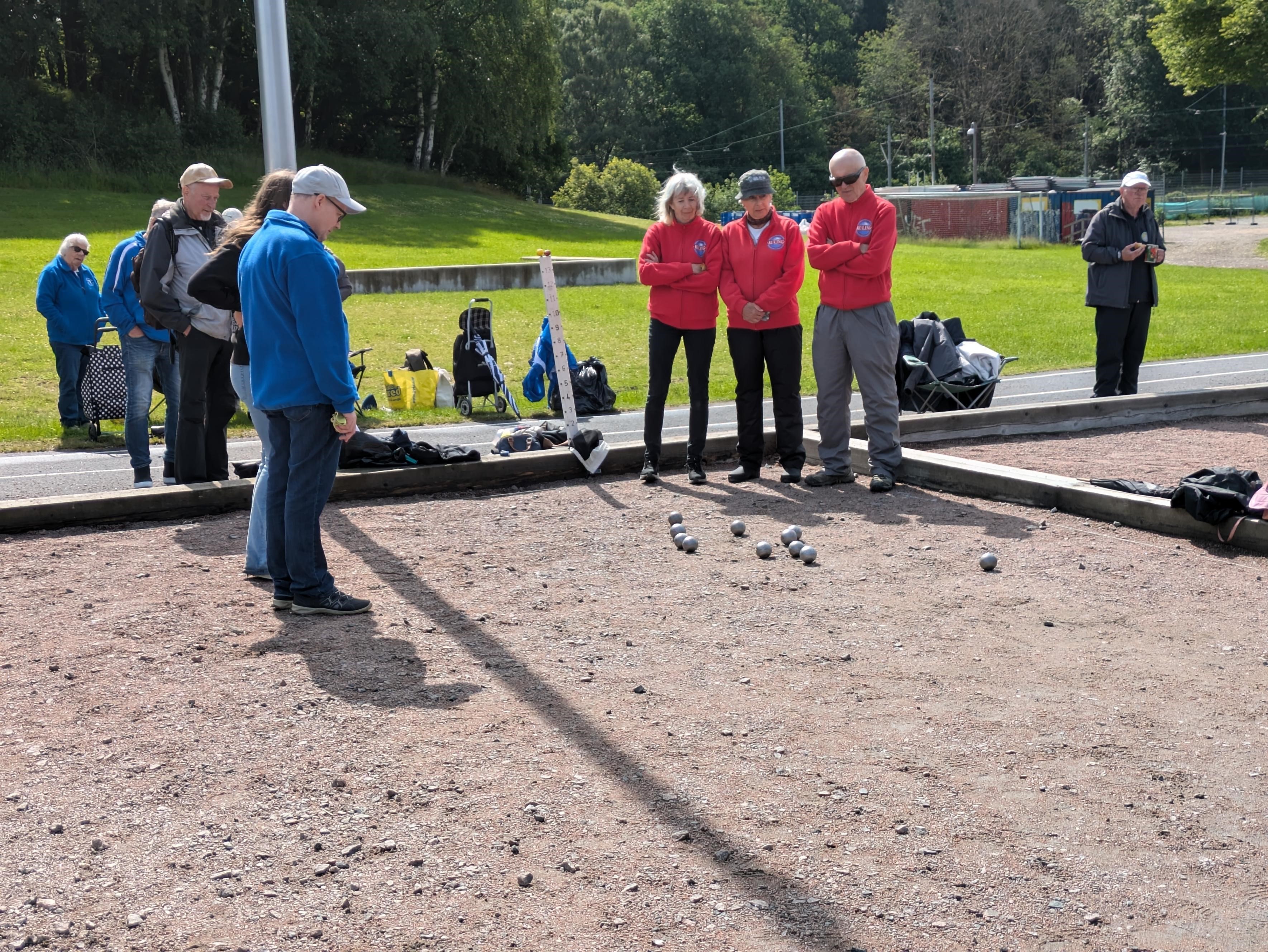 Ett gäng personer står vid en grusbana och spelar boule. Fyra personer i röda jackor tittar koncentrerat ner på kloten som ligger utspridda på marken. En person i blå jacka står vid sidan av och betraktar spelet. I bakgrunden syns fler personer, gräsytor och träd. Vädret är klart men kyligt.