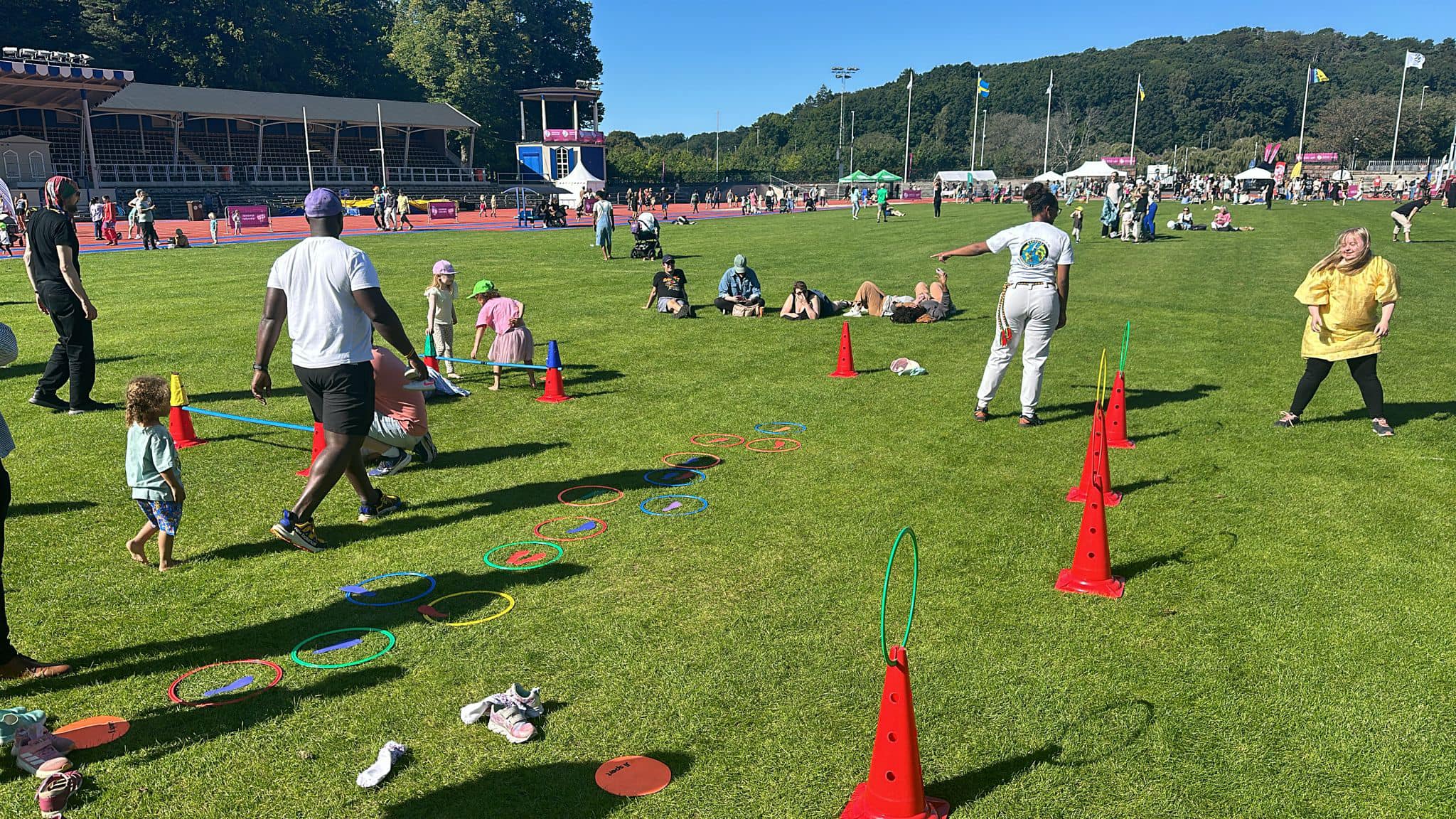Personer i olika åldrar deltar i lekfulla aktiviteter på en stor gräsmatta i Slottsskogsvallen. Rödvita koner, rockringar och färgglada markeringar används som hinderbana. Några vuxna vägleder barnen i banan, medan andra sitter i gräset. Det är soligt och många människor syns i bakgrunden vid andra stationer.