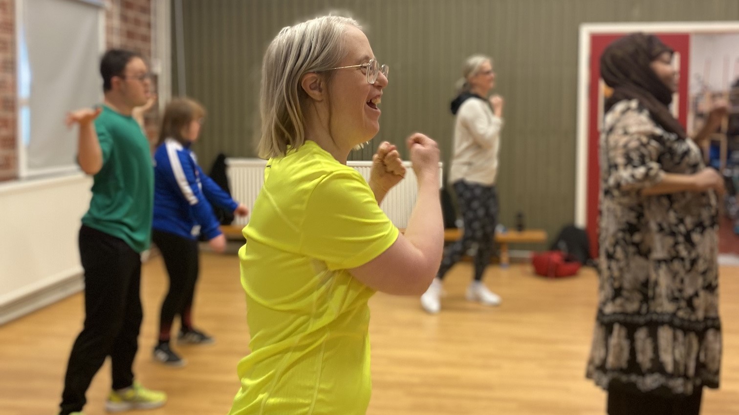 Ett gäng personer deltar i en gruppträning inomhus. I fokus står en kvinna med blont hår och glasögon i en gul t-shirt som ler stort och rör armarna uppåt. Övriga deltagare, i olika åldrar och kläder, rör sig i samma riktning. Atmosfären är energifylld och glädjefylld.