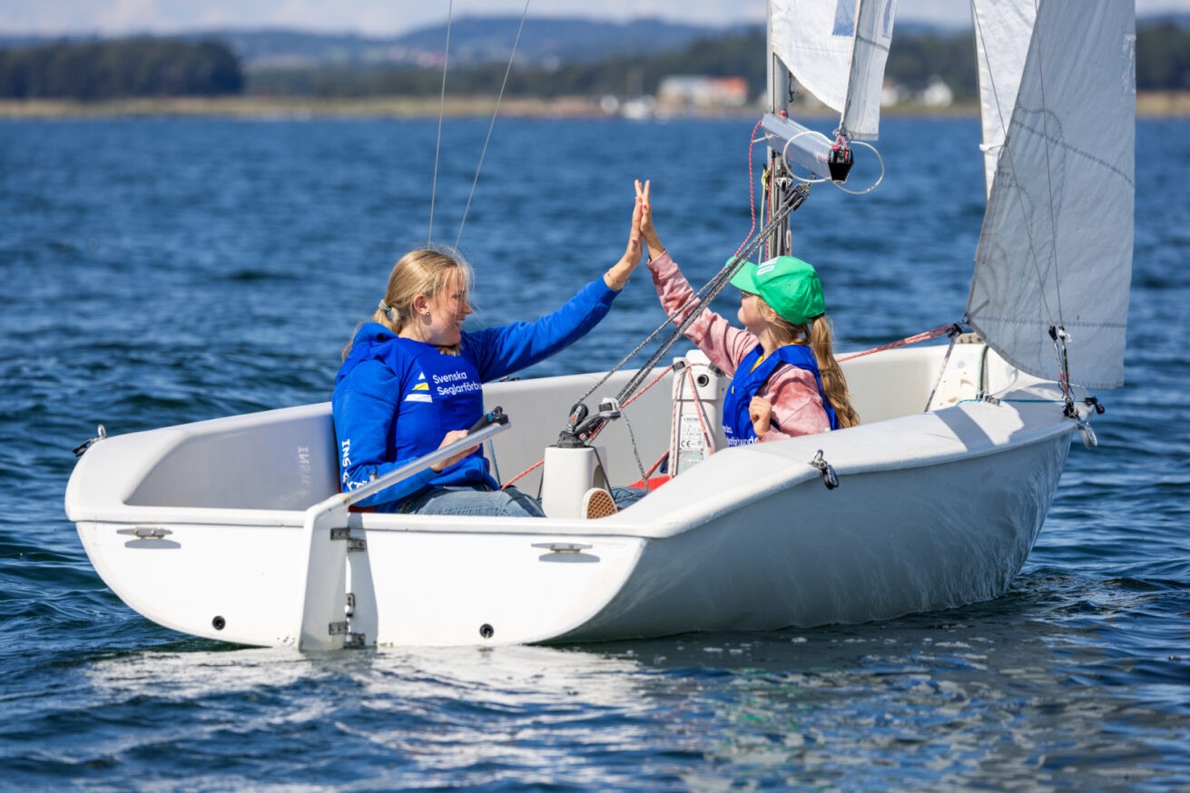 Två personer sitter i en liten vit segelbåt på öppet vatten. De bär blå flytvästar och ger varandra en high five. Den ena personen har en grön keps och rosa tröja, den andra har blå tröja med texten