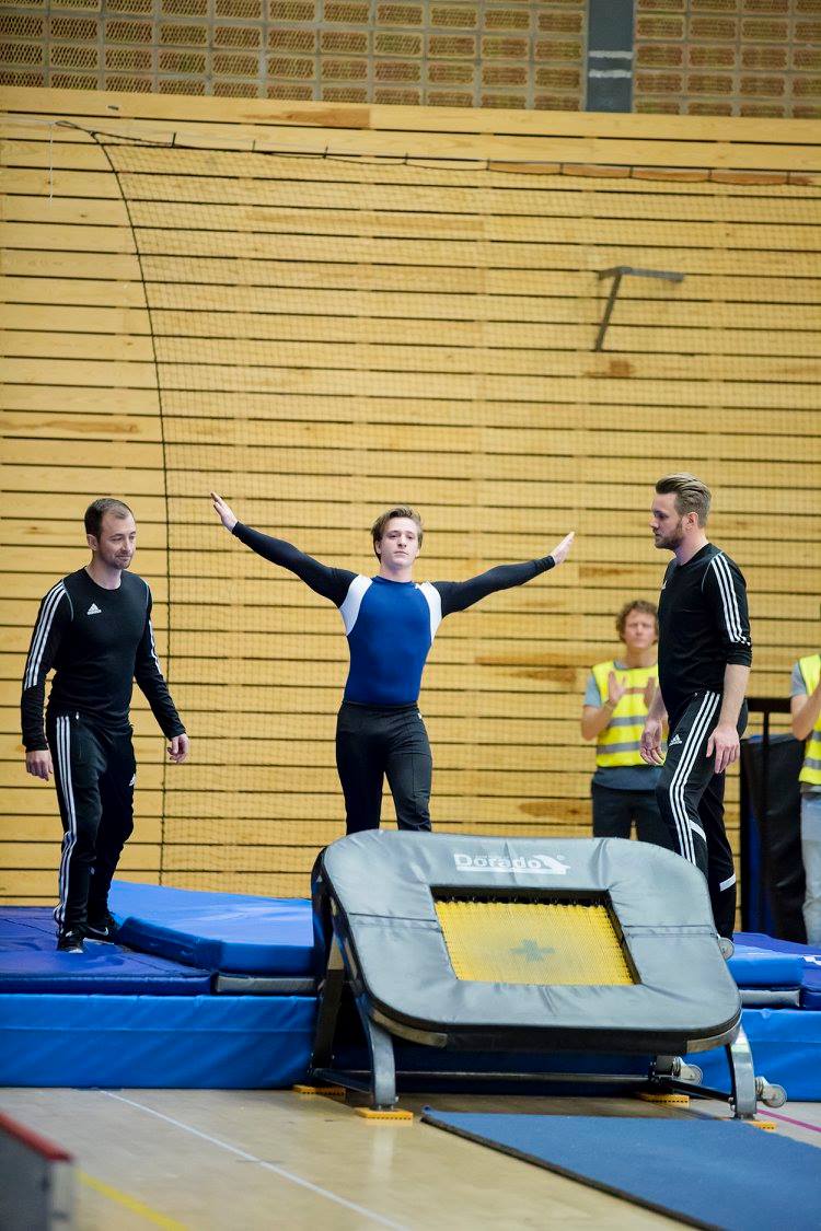 En person står med armarna utsträckta på en trampett i en gymnastikhall, redo att hoppa eller precis efter landning. Två ledare i svarta träningskläder står på varsin sida för att stötta. I bakgrunden syns personer i reflexvästar som verkar vara funktionärer.