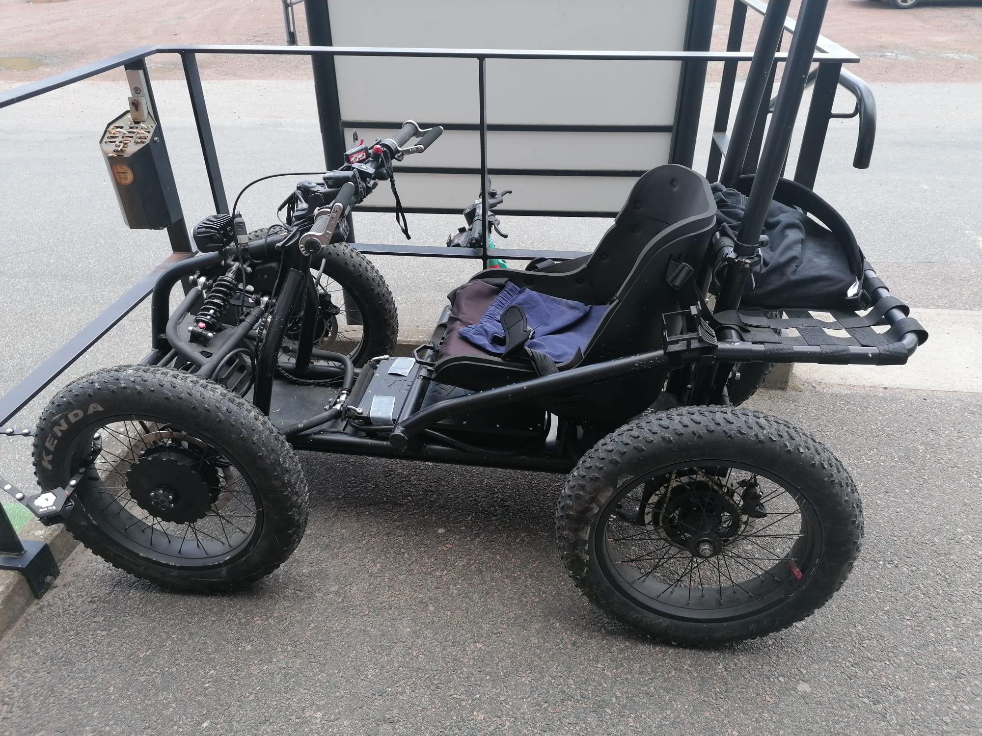 En svart, terränggående rullstol med fyra grova däck står parkerad på asfalt. Rullstolen har låg sittställning, fjädring framtill och handkontroller. Den är anpassad för att köras i ojämn terräng.