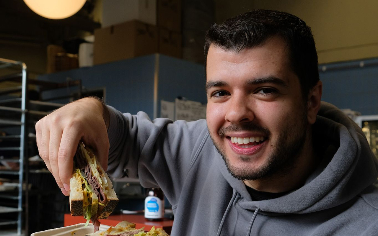 A young man with short dark hair and stubble smiles at the camera. He is holding up a large sandwich filled with various ingredients. He is wearing a grey hoodie and is sitting in what appears to be a kitchen or bakery, with shelves and boxes in the background.
