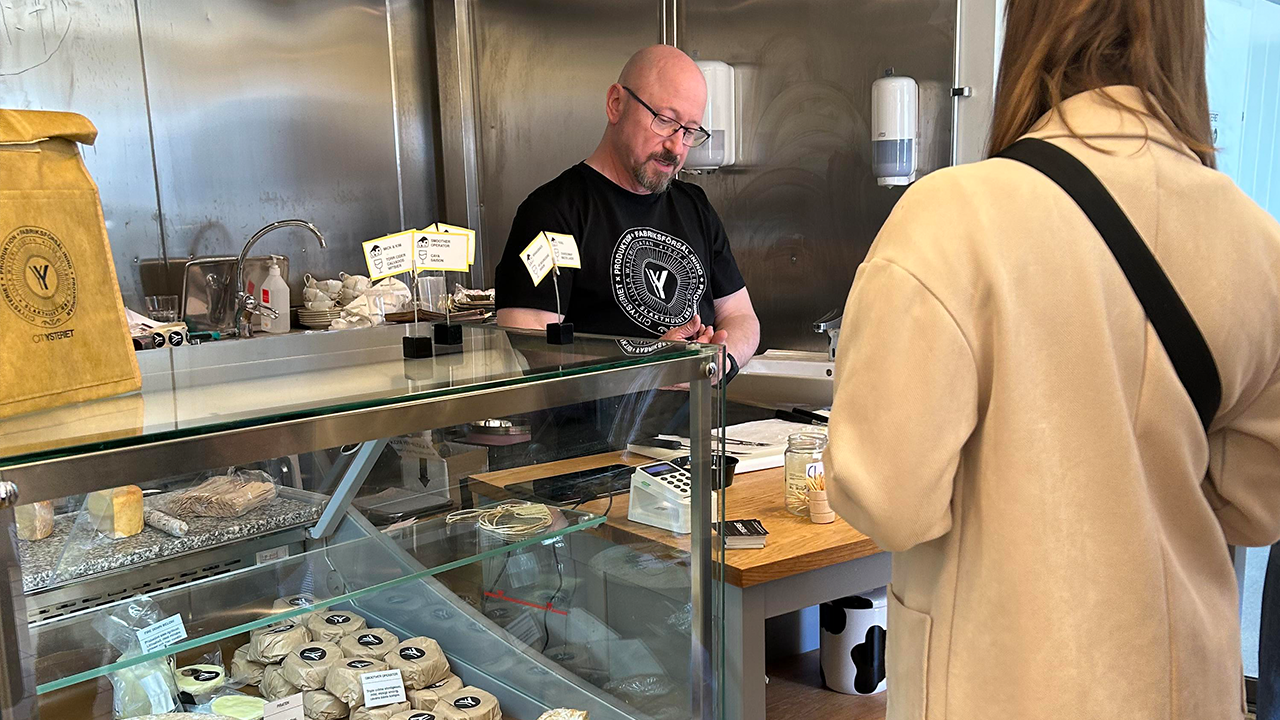 En man står bakom en ostdisk och pratar med en kund. I glasmontern framför honom ligger flera olika ostar framme.