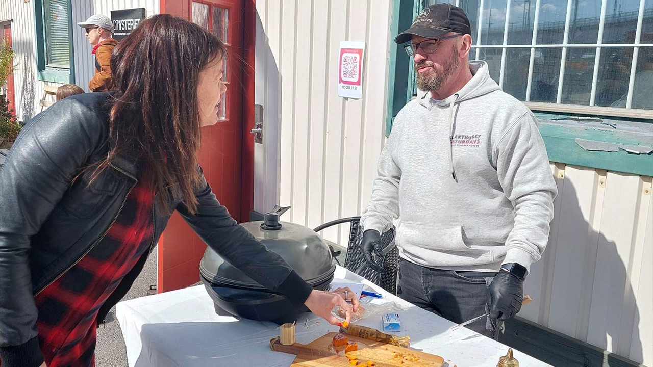 En kvinna sträcker sig fram för att ta en smakbit från ett bord. Bakom bordet står en man i grå hoodie och keps, redo att servera.