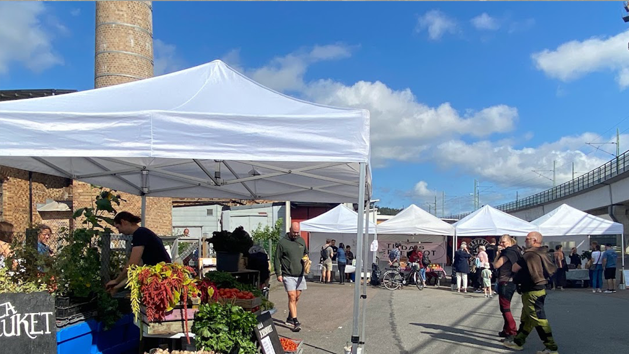 Flera vita tält står uppställda längs en gata vid en marknad. Människor går runt bland stånden under en blå himmel.
