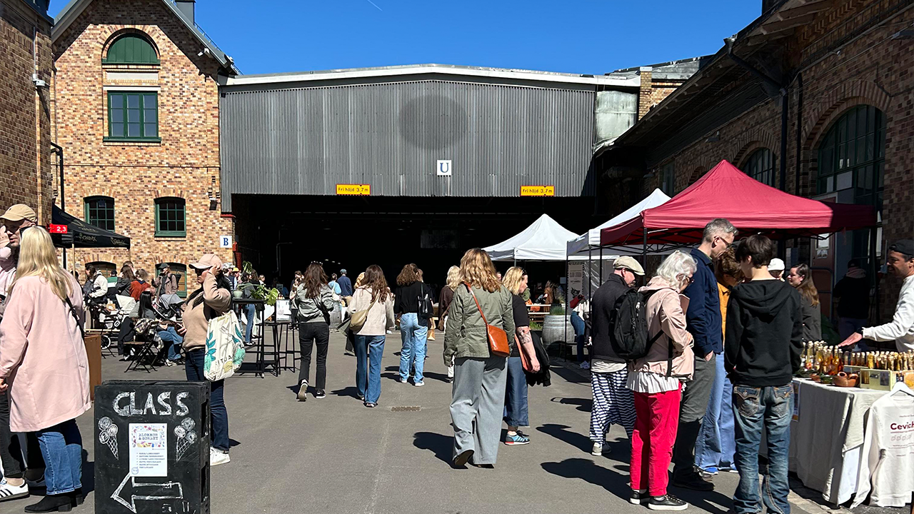 People walk and gather between market stalls with colorful tents. In the background, a large brick industrial building is visible.