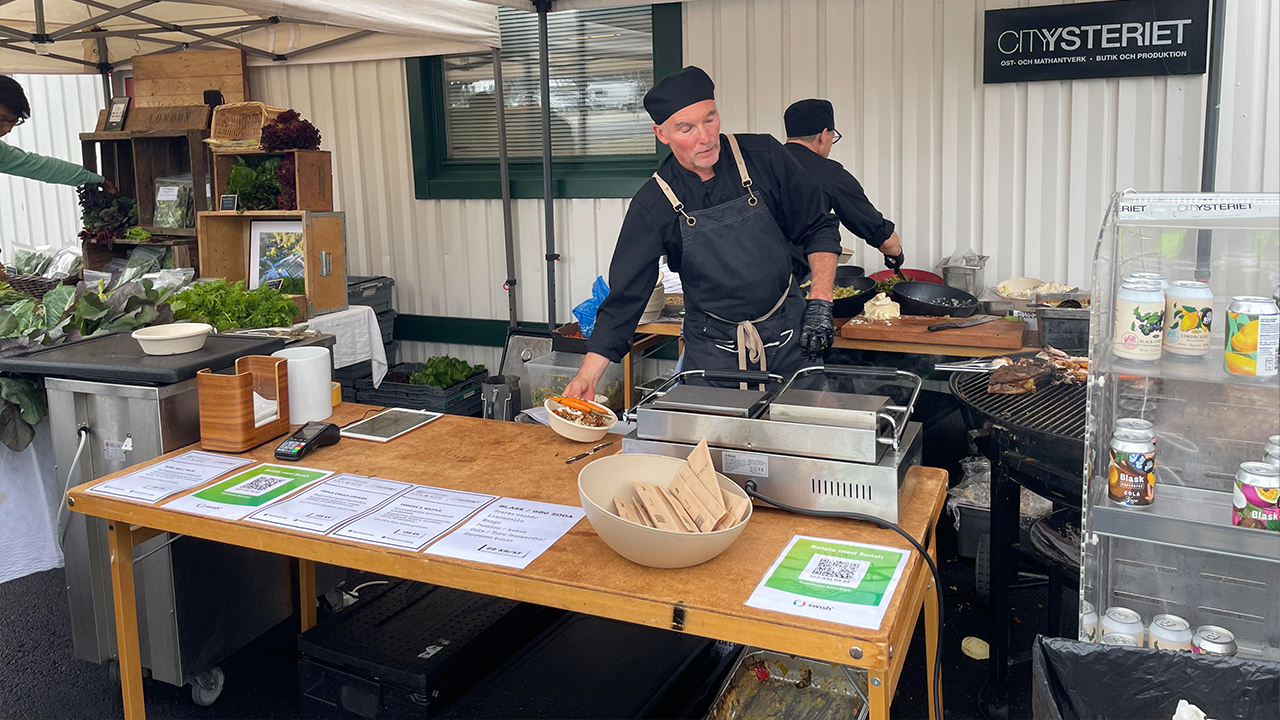 En man i svart kockkläder lagar mat vid ett marknadsstånd. På bordet framför ligger menyer, skålar och bestick.