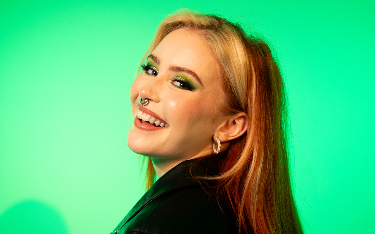 A woman is smiling broadly in front of a green background. She has orange-colored hair and a rock-inspired makeup look, with green eyeshadow and a septum piercing.
