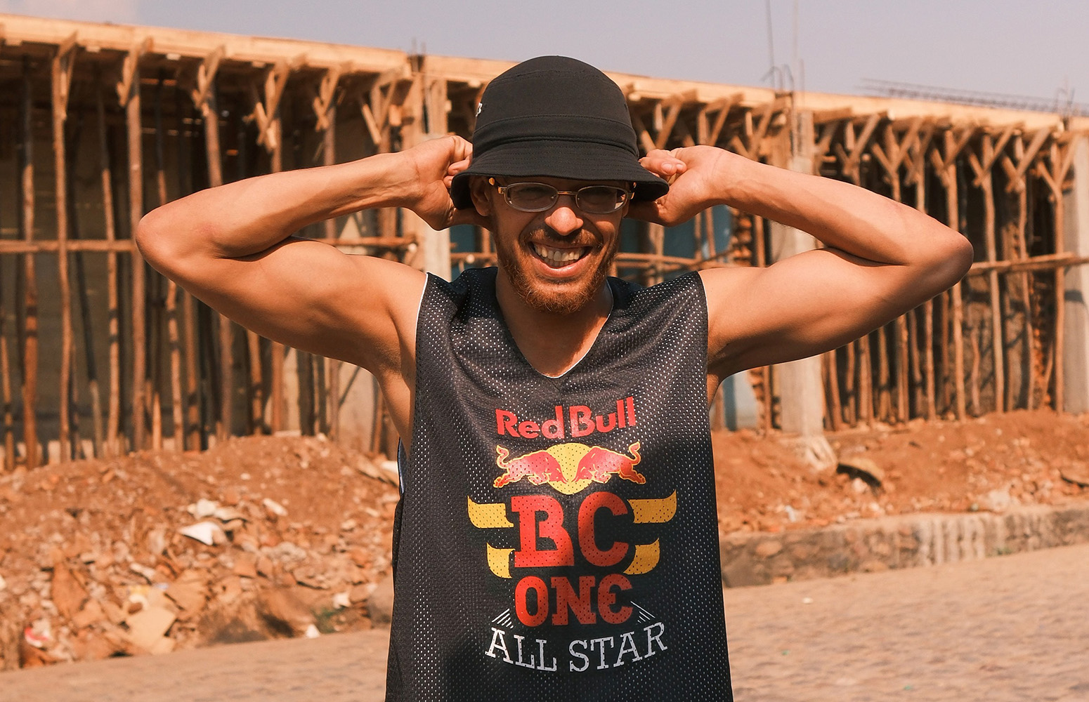 En ung man står utomhus framför en byggarbetsplats. Han ler stort, har solglasögon och en svart hatt. Han drar i hattens brätten med båda händerna och bär ett svart linne med texten ”Red Bull BC One All Star”.