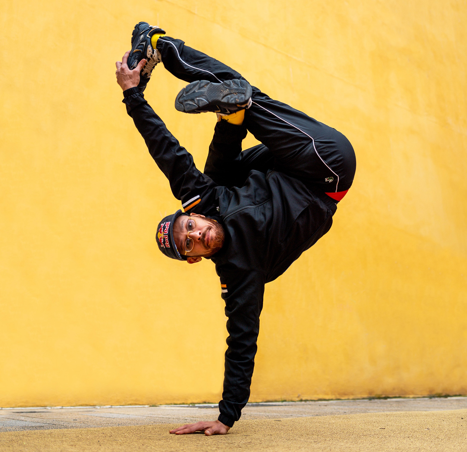 En ung man gör ett akrobatiskt dansmove mot en gul vägg. Han balanserar på ena handen med benen i luften i en avancerad breakdance-position. Han har svarta kläder, mössa och sneakers, och tittar mot kameran.