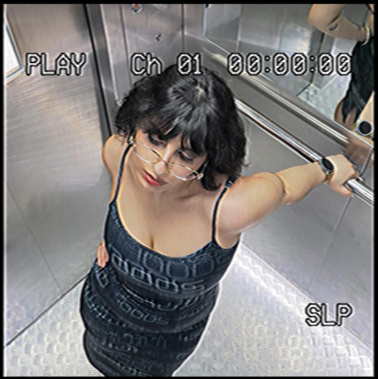 En person står i en hiss, fotograferad uppifrån. Bilden är stiliserad som en övervakningskamera med texten ”PLAY”, ”Ch 01” och tidskod i överkanten. Personen bär en svart klänning med tunna axelband och glasögon. Hen håller ena handen mot väggen och tittar något nedåt.