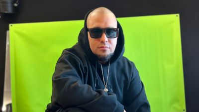 En person med rakat huvud och skägg sitter framför en grön bakgrund. Hen bär en svart huvtröja, svarta solglasögon och ett halsband med ett hängsmycke. Personen har armarna i kors och tittar rakt in i kameran med ett neutralt ansiktsuttryck. Bakgrunden är en grön duk uppsatt mot en mörk vägg.