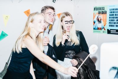 Unga personer står och ler in i en kamera framför en fotovägg klädd med flaggor i olika färger.