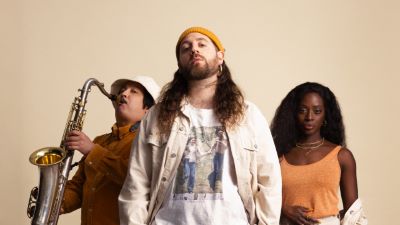 Tre personer: en som spelar saxofon, en man med långt hår och orange mössa, och en kvinna med mörkt långt hår.