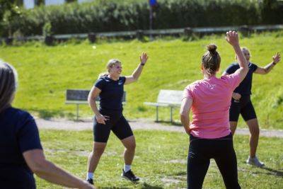 En person som har på sig sportkläder står mitt i en rörelse på en grön gräsmatta. Framför står flera personer och härmar rörelserna.