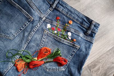 Bilden visar ett par blå jeans liggande på ett trägolv. Fokus ligger på bakfickan, där färgglada blommor är broderade så att de ser ut att växa upp ur fickan. Framför jeansen ligger flera nystan med broderigarn