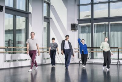 Fem dansare i ett synkroniserat danssteg. De står i en danssal med stora fönster.