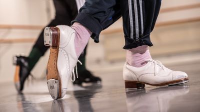 Närbild på två personer som steppar i en dansstudio. Fokus ligger på fötterna. Personen i förgrunden bär vita steppskor, rosa strumpor och mörkblå träningsbyxor med vita ränder.