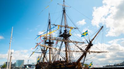 Ett stort skepp med flaggor och höga mastar. Står parkerat i en hamn med Göteborgs stadsmiljö i bakgrunden.