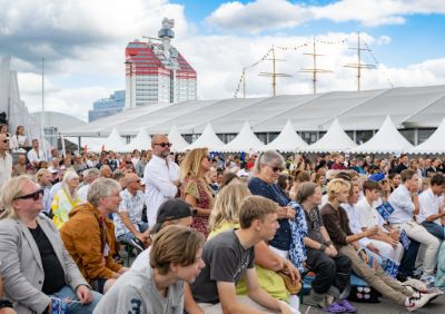 A large audience is seated outdoors in front of white tents, with Gothenburg’s iconic “Lipstick” building in the background.