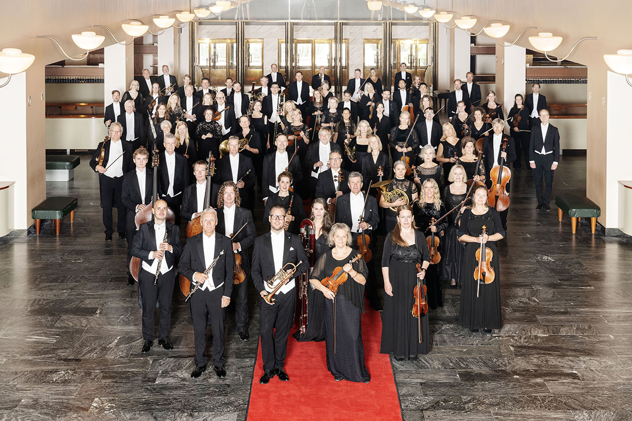 En stor orkester, en del i kavaj, en del i svarta klänningar står i ett elegant rum med en röd matta. Alla håller i varsitt insturment och kollar in i kameran.