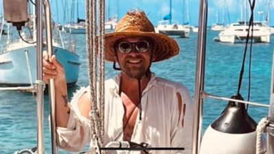 En man med stråhatt, vit skjorta och solglasögon sitter och ler på en segelbåt ute i turkosblått vatten. I bakgrunden syns flera segelbåtar.