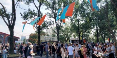 En stor folkmassa rör sig bland träd och stånd under en utomhusfestival. Färgglada tygband i orange och blått hänger i luften mellan träden. Många människor står i kö, sitter vid bord eller promenerar. I bakgrunden syns skyltar med texten ”Ö-festen” och matstånd.