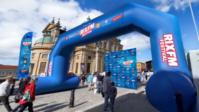 Ett stort blått uppblåsbart valv med texten RIX FM Festival står uppställt på ett torg framför en äldre kyrkobyggnad. Människor går in genom valvet mot ett område där en blå fotovägg med logotyper är uppställd.