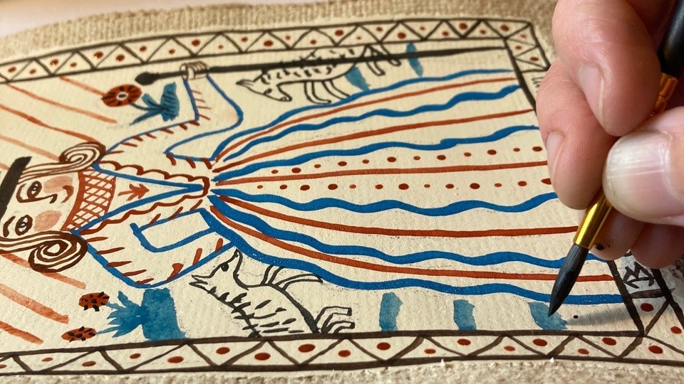 I bild syns en hand måla med en pensel på en papper. Målningen föreställer en figur med klänning.