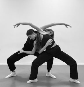En svartvit bild med två dansar som står i en danspose. De är klädda i svarta kläder, linne och byxor. Bakgrunden visar en kal vägg.