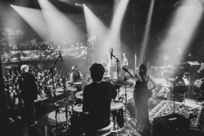 En svartvit bild visar ett liveuppträdande på en stor scen inför en entusiastisk publik. Bilden är tagen bakifrån scenen, där flera musiker syns spela – bland annat en trummis, en keyboardist och sångare.