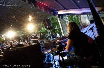 The silhouette of the drummer can be seen performing in front of an audience. In the background, stage lights and a forest environment are visible.