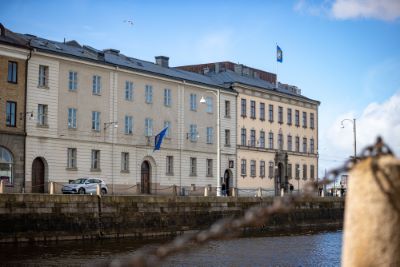 En ljus stenbyggnad med flaggor på fasaden och taket. Huset ligger vid vattnet under blå himmel. En kedja i förgrunden är suddigt synlig.