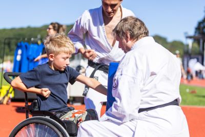 En pojke i rullstol tränar kampsport utomhus. Han slår mot en träningskudde som hålls av en instruktör i vit dräkt, medan en annan instruktör står bredvid.