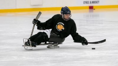 En paraishockeyspelare i svart tröja sitter i en kälke på isen och sträcker sig mot en puck med sin klubba. Spelaren bär hjälm och använder två korta klubbor med piggar i änden för att ta sig fram.