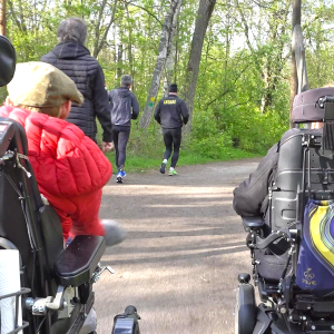 Två personer i elrullstol följer efter tre personer som joggar på en grusväg genom en grönskande skog. En av personerna i rullstol bär en röd jacka och keps, medan den andra har en svart mössa och jacka. Det är vår eller sommar, med gröna träd runt stigen.