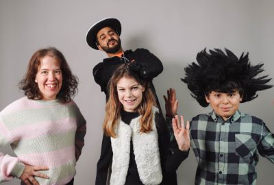 Fyra personer poserar framför en grå bakgrund. En man står längst bak med hatt och armarna i kors, medan tre barn och ungdomar ler mot kameran – en med randig tröja, en med vit pälsväst och en med rutig skjorta och stor svart peruk.