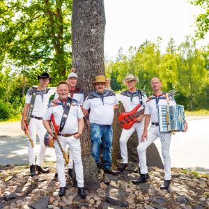 Six men are standing outdoors in a summery forest setting. They are dressed in light clothing, and some are holding instruments like a guitar and an accordion. The background shows green trees and sunshine.