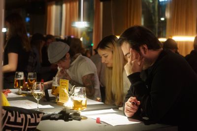 Personer sitter koncentrerat vid ett bord på en pub eller restaurang, med pennor i händerna och quizpapper framför sig. Ölglas och levande ljus står på borden. Miljön är varm och stämningsfull.