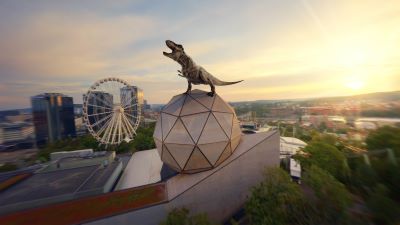 En digitalt skapad T. Rex står på Wisdome-kupolen vid Universeum i Göteborg. I bakgrunden syns stadens silhuett och Lisebergshjulet i solnedgång.