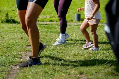 Närbild på ben och fötter av flera personer som tränar utomhus på gräs. En vuxen och ett barn deltar tillsammans, barnet har sandaler och shorts, medan de vuxna bär träningskläder och joggingskor.