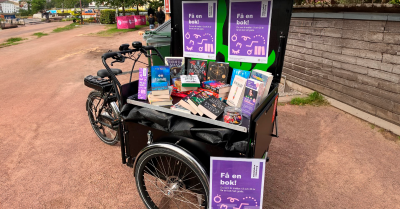 En lådcykel fylld med böcker står parkerad utomhus. På cykeln sitter skyltar med texten