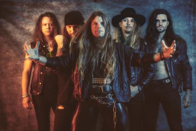 Ett hårdrocksband poserar framför en mörk, studioliknande bakgrund. I centrum står en man med långt, blont hår, skinnbyxor och utsträckta armar, som ser direkt in i kameran med intensiv blick. Runt honom står fyra andra bandmedlemmar, alla med långt hår och klädda i svarta rockkläder. Stämningen är allvarlig och kraftfull.