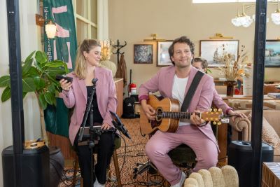 A woman and a man wearing matching pink blazers sit side by side. The man plays guitar while the woman holds a microphone and a shaker. They look happy.
