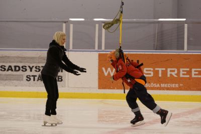 Ett barn i hjälm och sele svävar ovanför isen i en ishall, fastspänt i en sele från taket. En vuxen på skridskor sträcker ut armarna mot barnet för att ta emot det.