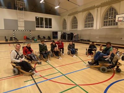 En grupp personer sitter i en gymnastiksal på specialbyggda, låga rullstolar med stora hjul. De sitter i en cirkel och ser glada ut – flera av dem vinkar eller gör tummen upp. Hallen är ljus med stora fönster och färgade linjer på golvet.