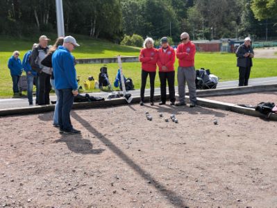 Ett gäng personer står vid en grusbana och spelar boule. Fyra personer i röda jackor tittar koncentrerat ner på kloten som ligger utspridda på marken. En person i blå jacka står vid sidan av och betraktar spelet. I bakgrunden syns fler personer, gräsytor och träd. Vädret är klart men kyligt.