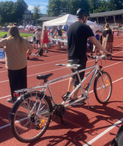 En person med svart t-shirt och cykelhjälm står på en röd löparbana och håller i en silverfärgad tandemcykel. I bakgrunden syns fler personer, tält och aktiviteter. Det är soligt och ser ut att vara en festival eller ett event.