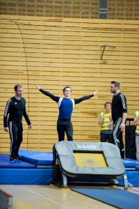 En person står med armarna utsträckta på en trampett i en gymnastikhall, redo att hoppa eller precis efter landning. Två ledare i svarta träningskläder står på varsin sida för att stötta. I bakgrunden syns personer i reflexvästar som verkar vara funktionärer.